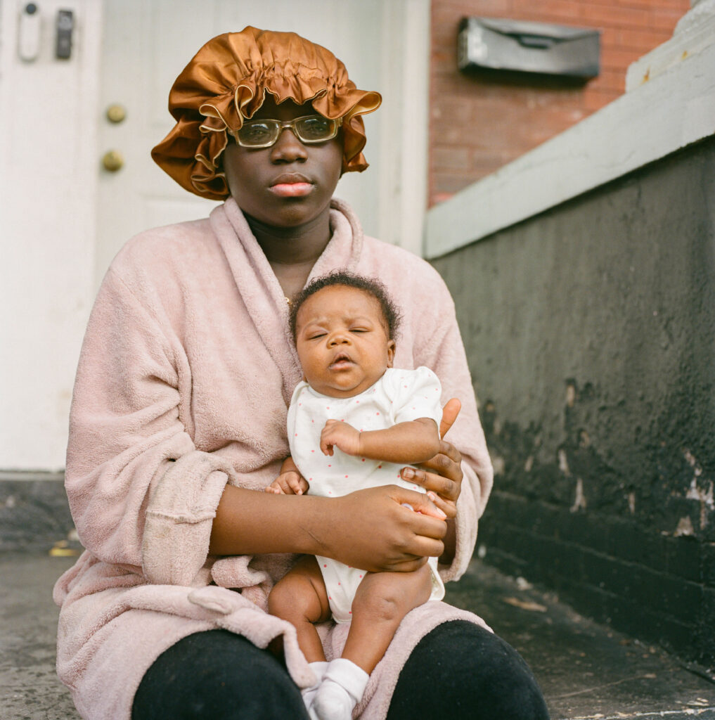 A young woman in postpartum holds her baby as she watches neighbors from her doorway. Unable to afford a hotel or relocation, she remains in her storm-damaged home, which now bears deep cracks. She asked to remain anonymous.