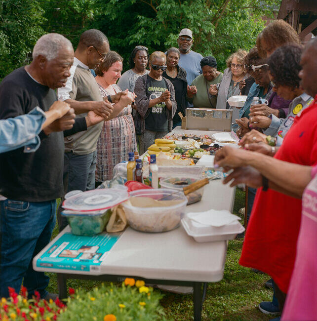 Members of the Lewis Place Historic Preservation community bow their heads in prayer before sharing a meal. They offer thanks for the food and lift prayers for social and environmental justice, asking God to bring healing and equity to their neighborhood.