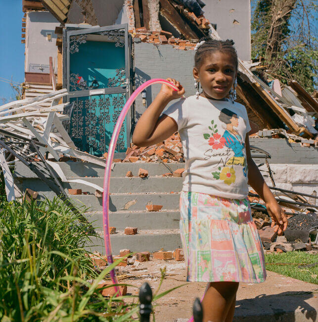 A girl in front of her defrayed house playing with her pink hula hoop.